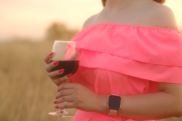 Unrecognizable woman holding glass of red wine standing in nature on sunset Body part of female in bright dress with alcohol in countryside