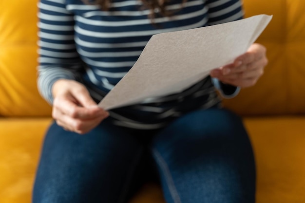 An unrecognizable woman sits on a yellow sofa and holds a blank sheet of paper in her hands your ad woman shows poster canvas white sheet of paper