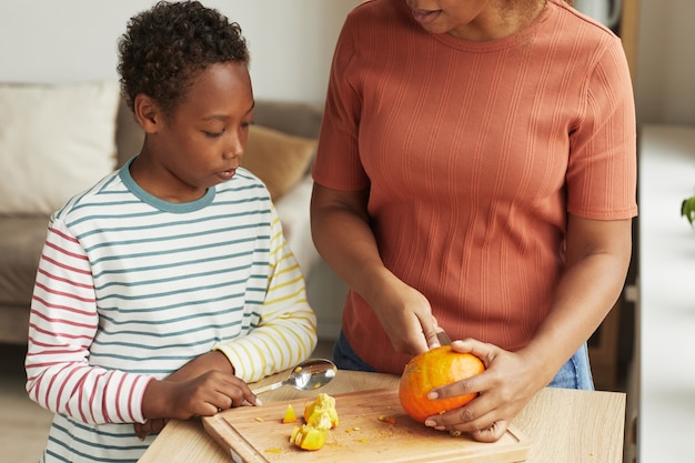 Unrecognizable woman standing at kitchen counter demonstrating her son how to carve pumpkin for Halloween