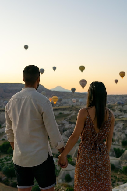 Unrecognizable young couple holding hands in the foreground looking at the typical hot air balloons of the cappadocia area flying at sunrise