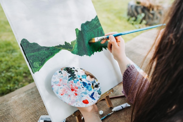 Unrecognizable young female artist painting her own mountain scenery with oil on a canvas. Amateur