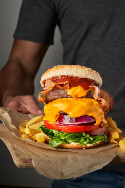 Unrecognizable young man holding a wooden table with tasty fast burger and fries on grey background