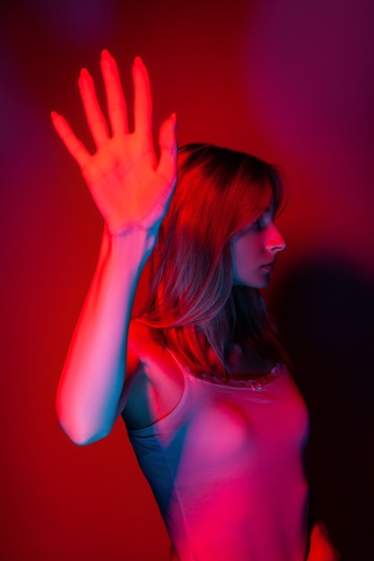 Unrecognizable young slim lady in light top covering face with hand while standing in dark red room