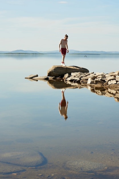 Photo unrecognizable young topless man walking on stones on background of picturesque calm lake and blue sky on summer afternoon