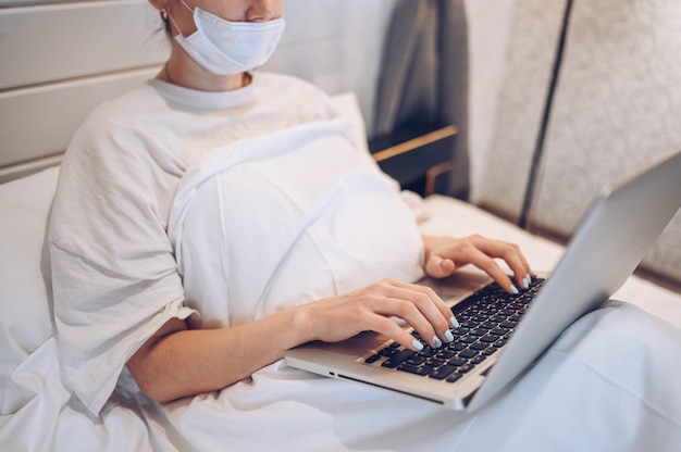 Unrecognizable young woman in face medicine mask  working on a laptop in bedroom during coronavirus isolation home quarantine. Covid-19 pandemic Corona virus. Distance online work from home concept.