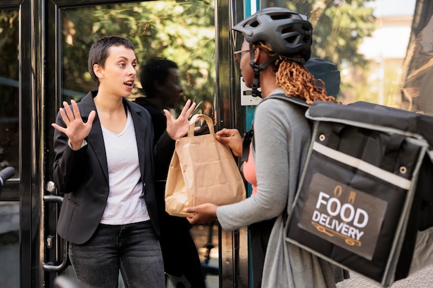 Unsatisfied client refuse taking late food order, delivery delay, courier and angry customer. Woman giving impulsive furious employee takeaway meal near office building outdoors