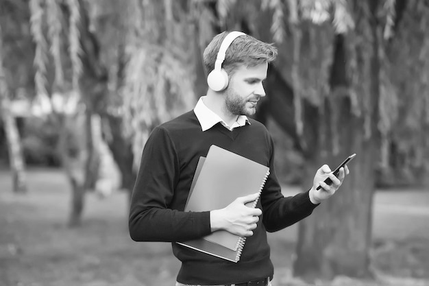 Unshaven guy holding notebook and listening music in earphones from smartphone education