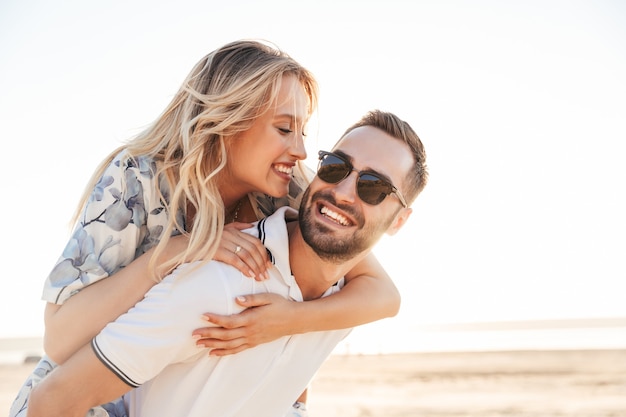 unshaven happy man wearing sunglasses smiling and giving piggyback ride seductive woman while walking on sunny beach