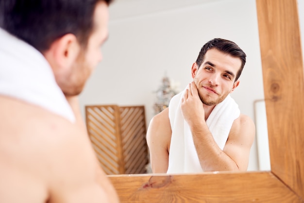 Unshaven undressed brunet with white towel on his neck standing near mirror