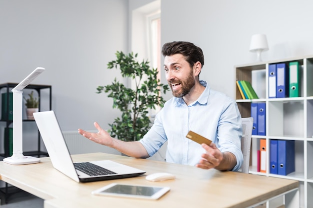 Unsuccessful online purchase Unsuccessful deal Upset young man office worker holding a credit card holding his head in shock Sitting at a desk at a laptop