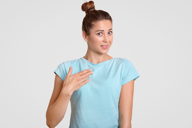 Unsure woman keeps hand on chest, asking why she was chosen, stands against white wall, wears casul t shirt, isolated over white. People doubt and shyness concept.