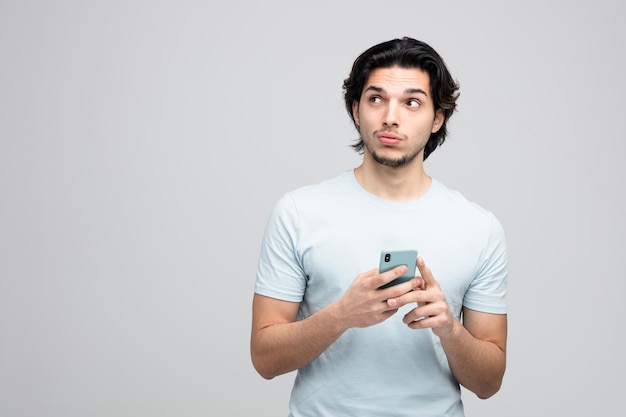 unsure young handsome man holding mobile phone looking at side isolated on white background with copy space