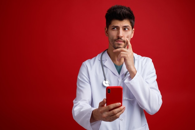 Unsure young male doctor wearing medical uniform and stethoscope around his neck holding mobile phone keeping hand on chin looking at side isolated on red background with copy space