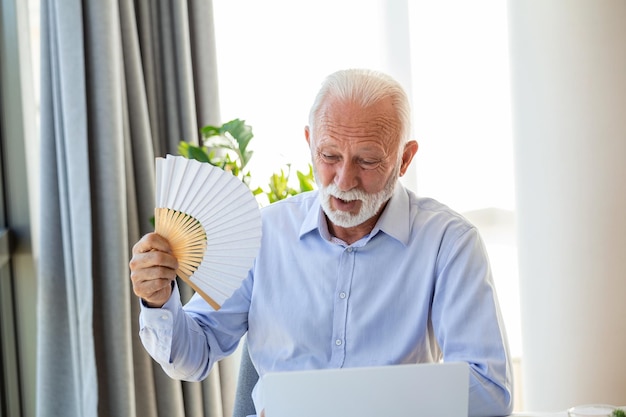 Unwell senior business man sit on desk work on laptop wave with hand fan Overheated man worker use waver suffer from heatstroke in office struggle with no AC at workplace