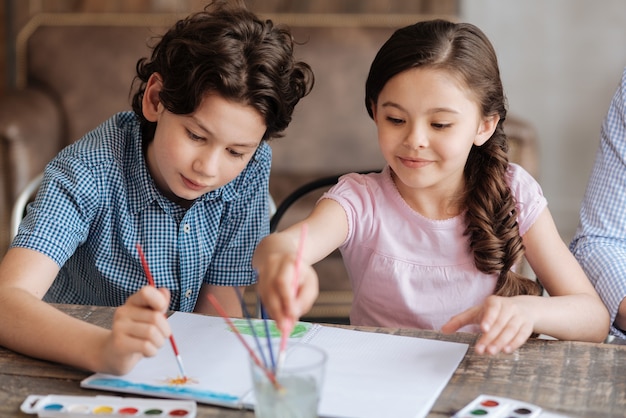 Upbeat adorable children painting a watercolor picture together with the boy depicting the sun and his sister washing a brush in the glass