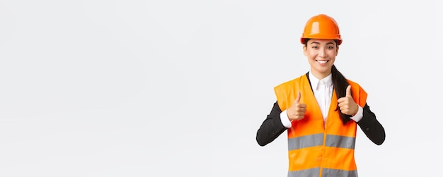 Upbeat pleased asian female architect giving permission proud with result standing in safety helmet and reflective jacket showing thumbsup in approval guarantee best construction quality