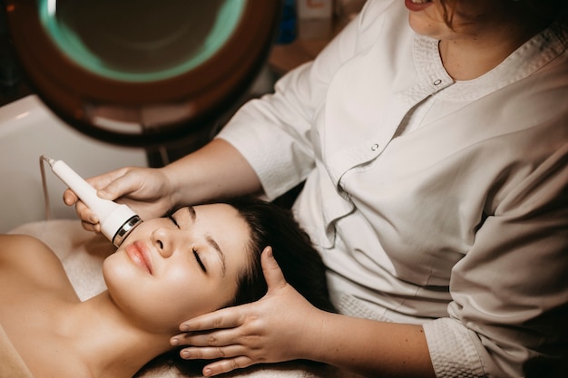 Upper view of a beautiful female having electroporation procedure on face in a spa resort .