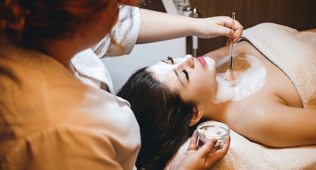 Upper view of a charming female with dark hair leanin with closed eyes while having a skin care white mask in a spa resort.