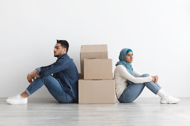 Upset arab family sitting separated by boxes on floor in empty apartment, having problem with relocating, white wall background, side view, copy space. Depressed middle-eastern couple at new home