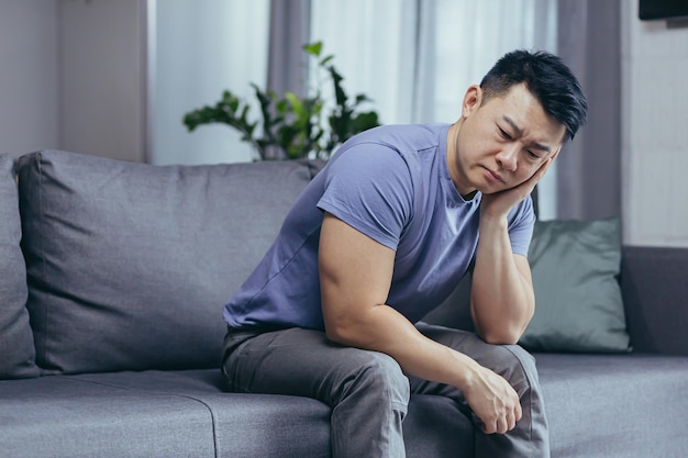 Upset Asian man at home tired and depressed sitting on the couch in the living room