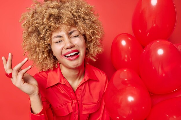 Upset curly haired woman with miserable expression keeps hand raised has leaked makeup wears shirt holds inflated heart balloons dressed in shirt isolated over red background Celebration concept