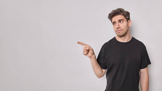 Upset discontent European adult man points away with index finger shows something unpleasant doesnt like this advertisement dressed in black t shirt isolated over white background. Mock up space