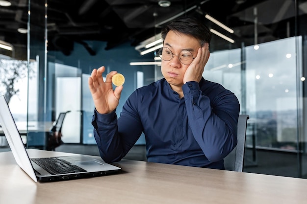 Upset and dissatisfied young man asian businessman holds a bitcoin in his hands looks at it sadly