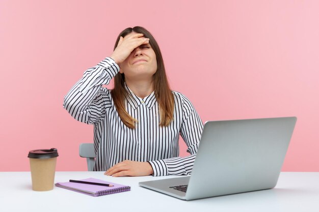 Photo upset frustrated woman office worker in striped shirt touching face with hand making facepalm gesture sitting at laptop difficulties at work mistake indoor studio shot isolated on pink background