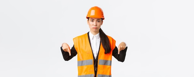 Upset and gloomy asian female construction manager in safety helmet and reflective jacket pointing fingers down and pouting disappointed showing bad news white background