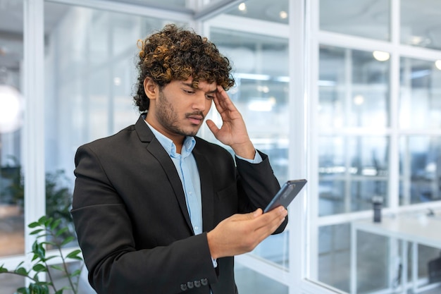 Upset hispanic man in office at work reading bad news from phone online businessman in shirt near