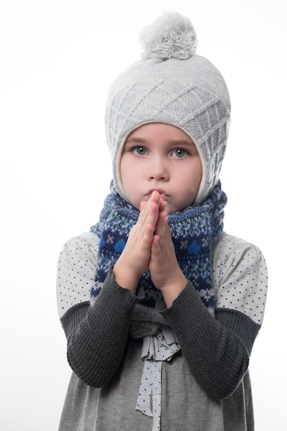 Upset little girl in a gray hat wrapped in a scarf on a white background waiting for the cold winter