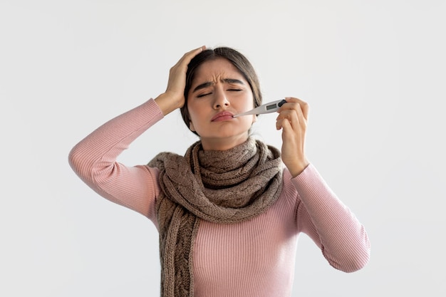 Upset millennial indian lady in scarf measures temperature with thermometer in mouth and suffers from headache