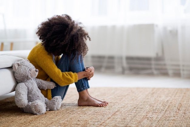 Upset preteen black girl sitting on floor with teddy bear