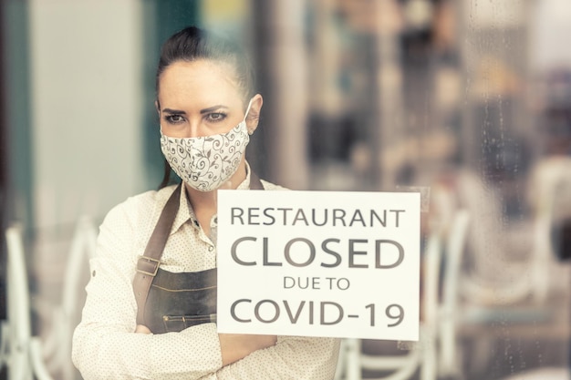 Upset restaurant owner stands with arms crossed on her chest next to a sign saying restaurant closed due to covid19