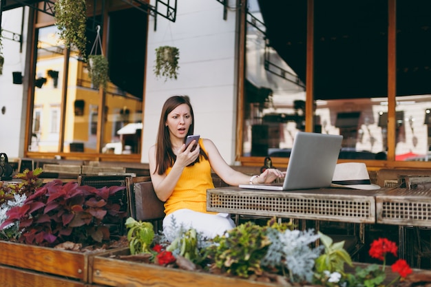 Upset sad girl in outdoors street cafe sitting with laptop pc computer, looking on mobile phone texting message sms, disturb of problem, in restaurant during free time