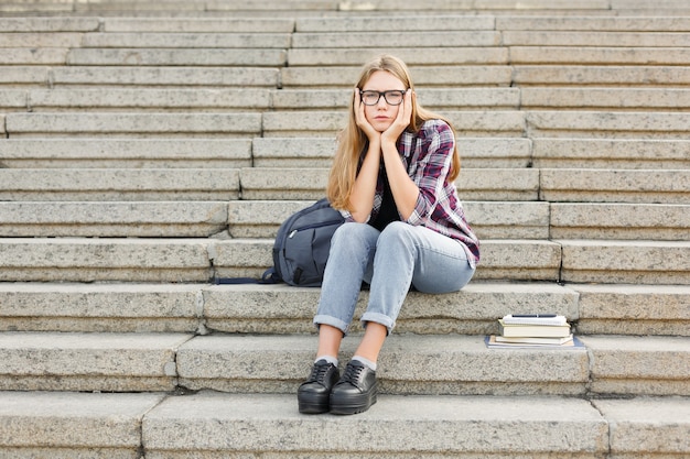 Upset student girl sitting on stairs outdoors in university campus. Woman raised hands to her head. Education concept, copy space