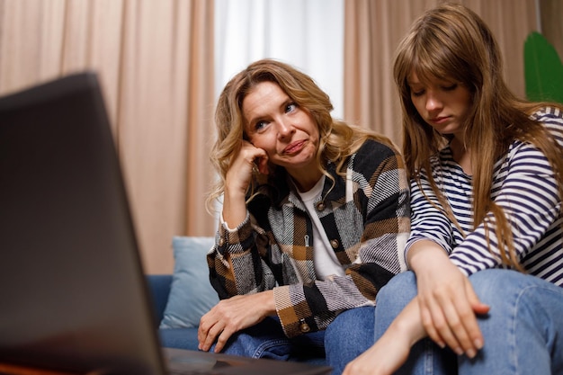 Upset women of different ages sitting on sofa in living room looking at laptop