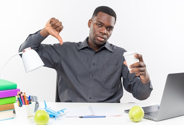 Upset young afro-american student sitting at desk with school tools holding paper cup and thumbing down 