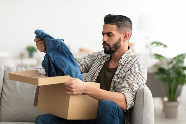 Upset young Arab guy unboxing carton package receiving new clothes unhappy about delivered item at home