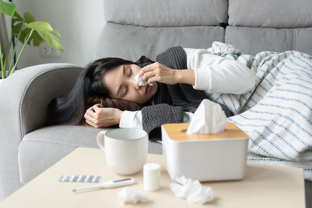 Upset young Asian woman sleeping on couch covered with blanket freezing blowing running nose got fever caught cold sneezing in tissue sick female having influenza symptoms sleep at home Flu concept