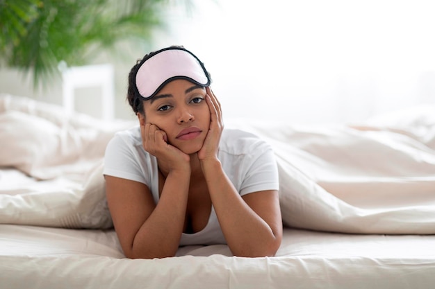 Upset young black woman wearing sleeping mask lying in bed at home