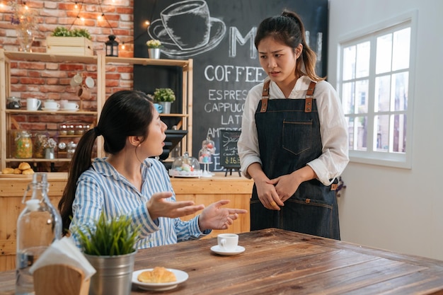 Upset young businesswoman complain about cup of hot coffee. woman customer hold it in hands and point to order while look at waitress. lady client complaining meal to waitress in cafe store.