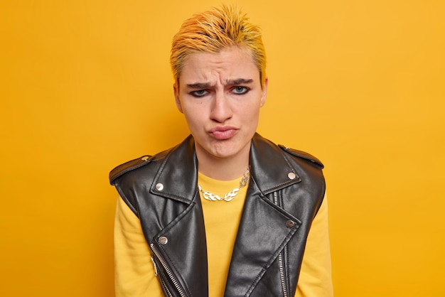 Upset young hipster girl with makeup trendy hairsyle dressed in casual jumper and black leather vest has displeased expression dissatisfied with something isolated on bright yellow