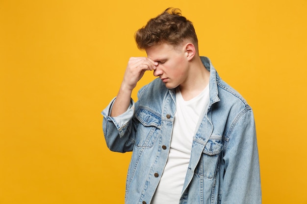 Upset young man in denim casual clothes with lowered head keeping eyes closed putting hand on nose isolated on yellow orange background. People sincere emotions, lifestyle concept. Mock up copy space.