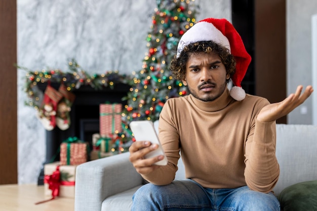 Upset young man sitting on sofa at home in red santa hat hispanic looking disappointed at camera