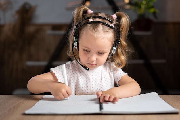 Upset young student in headset sitting at the table at home schoolgirl little child draws with a pen in a notebook