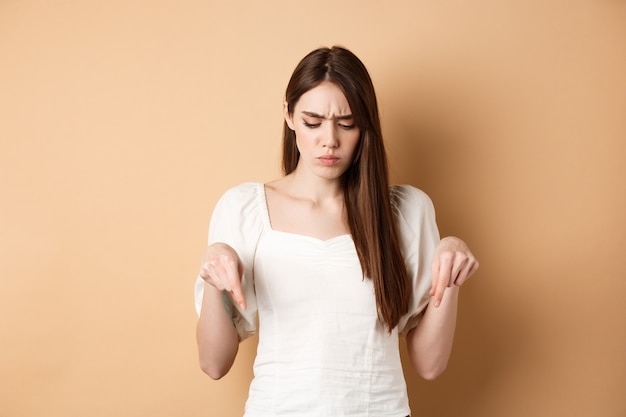 Upset young woman frowning and looking down pointing at bad thing standing confused and displeased o...