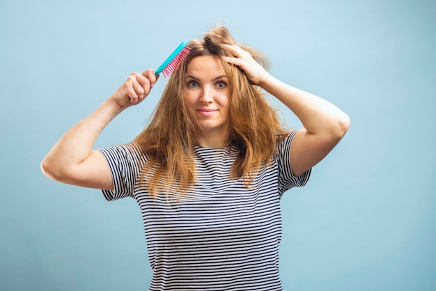 Upset young woman looking with shock at her damaged hair on blue background