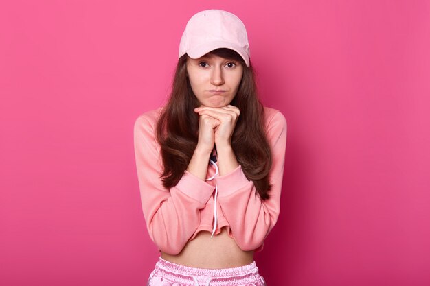 upset young woman with dissatisfied expression, keeps hand under chin, looks at camera with pouty lips, displeased to stay at home at weekend, wears stylish short hoodie. Teen concept.