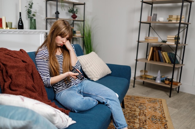 Upset young woman with TV remote control in hands sitting on sofa